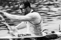 Dynamisches Schwarz-weiß-Foto von Max Lemke, dem dreifachen Olympiasieger, während eines Kanurennen. Der Athlet trägt ein weißes Trikot mit der Aufschrift 'Germany' und eine Sonnenbrille. Wassertropfen und Bewegungsunschärfe im Bild fangen die explosive Kraft und Intensität des Sports ein. Die kontrastreiche Aufnahme zeigt ihn während eines kraftvollen Paddelschlags.